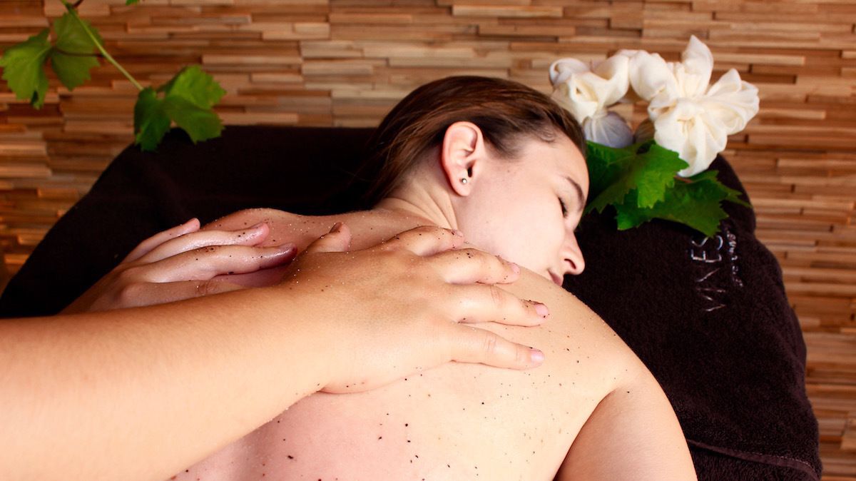 Une personne reçoit un massage, les mains de la masseuse étant posées sur son dos. L’arrière-plan est constitué d’un mur en bois, d’une serviette noire et de fleurs blanches.