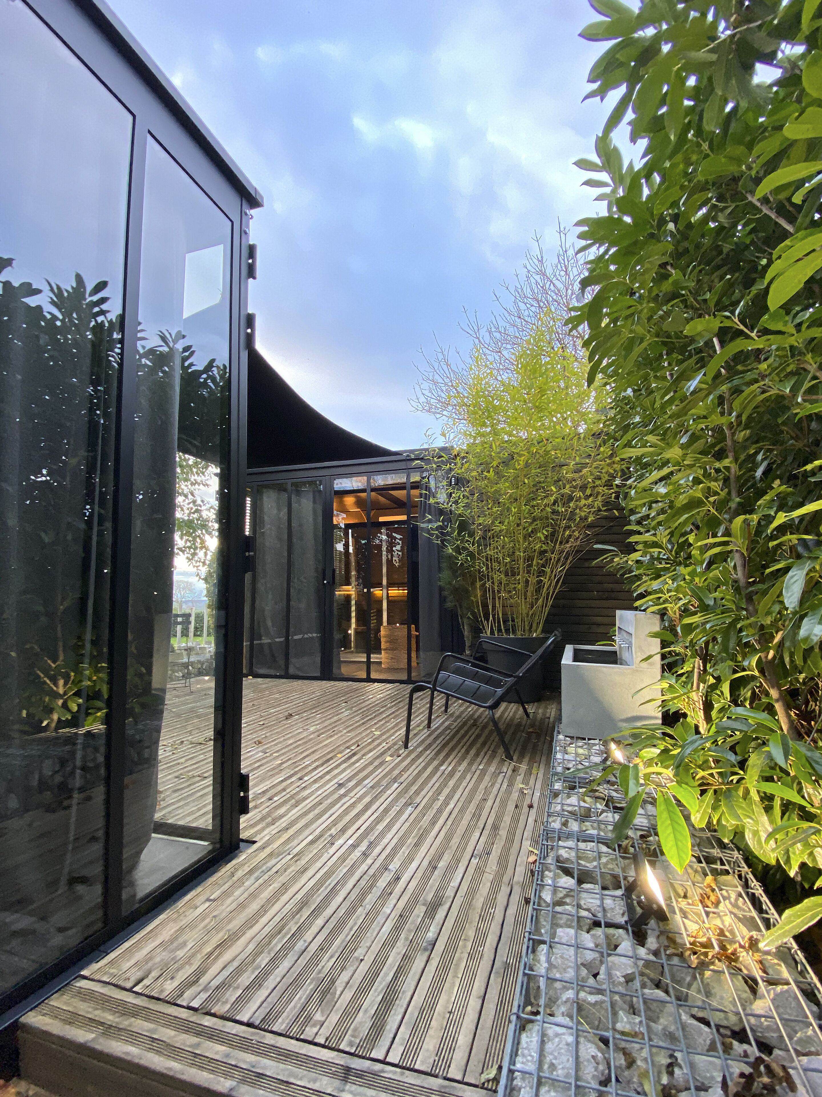 A modern outdoor space with a wooden deck, a glass door entrance, a seating area, and a bamboo plant.