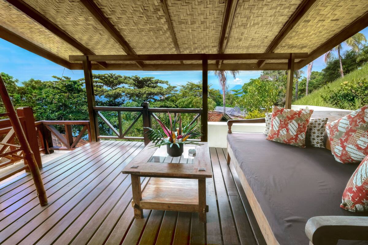 A wooden deck with a thatched roof, a sofa with patterned cushions, and a small table with a potted plant.