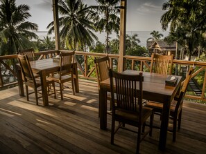 Una terraza de madera con una mesa y sillas, con vistas a un paisaje tropical con palmeras y al mar.