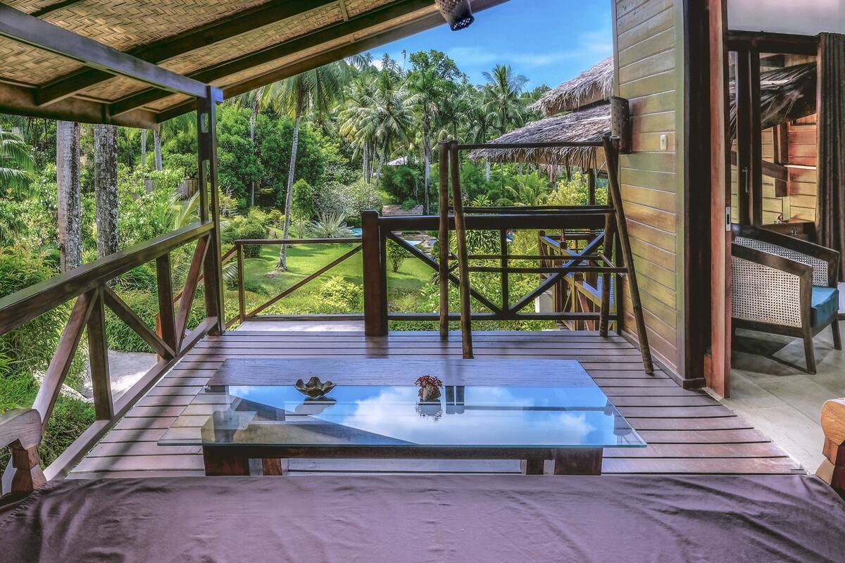 A wooden deck with a glass table and chairs, overlooking a lush garden with palm trees.