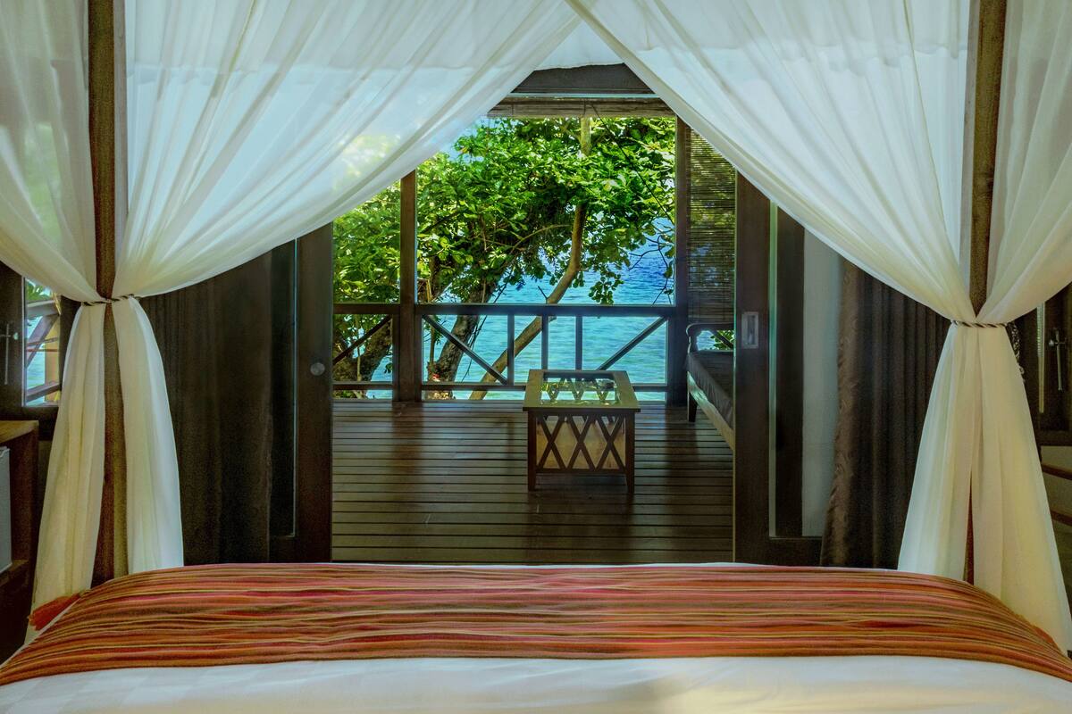 A bed with a striped bedspread, a wooden deck with a table, and a view of the sea and trees through a canopy.