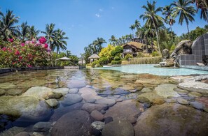 Una piscina cristalina con rocas, rodeada de palmeras y un edificio con techo de paja.