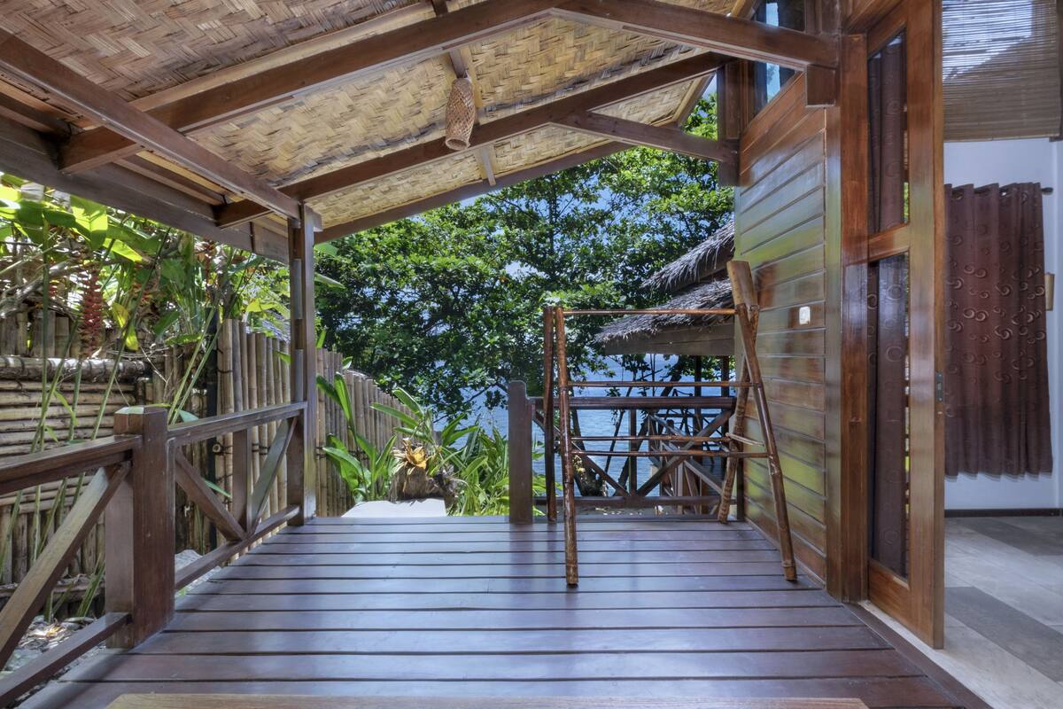 A wooden deck with a thatched roof, leading to a wooden gate and a ladder.