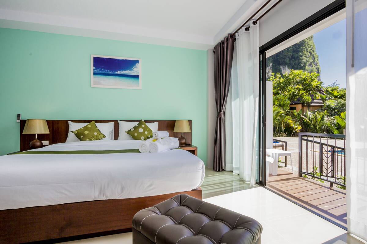 A hotel room with a large bed, two bedside lamps, a green pillow, a wooden headboard, and a view of a balcony with outdoor furniture.