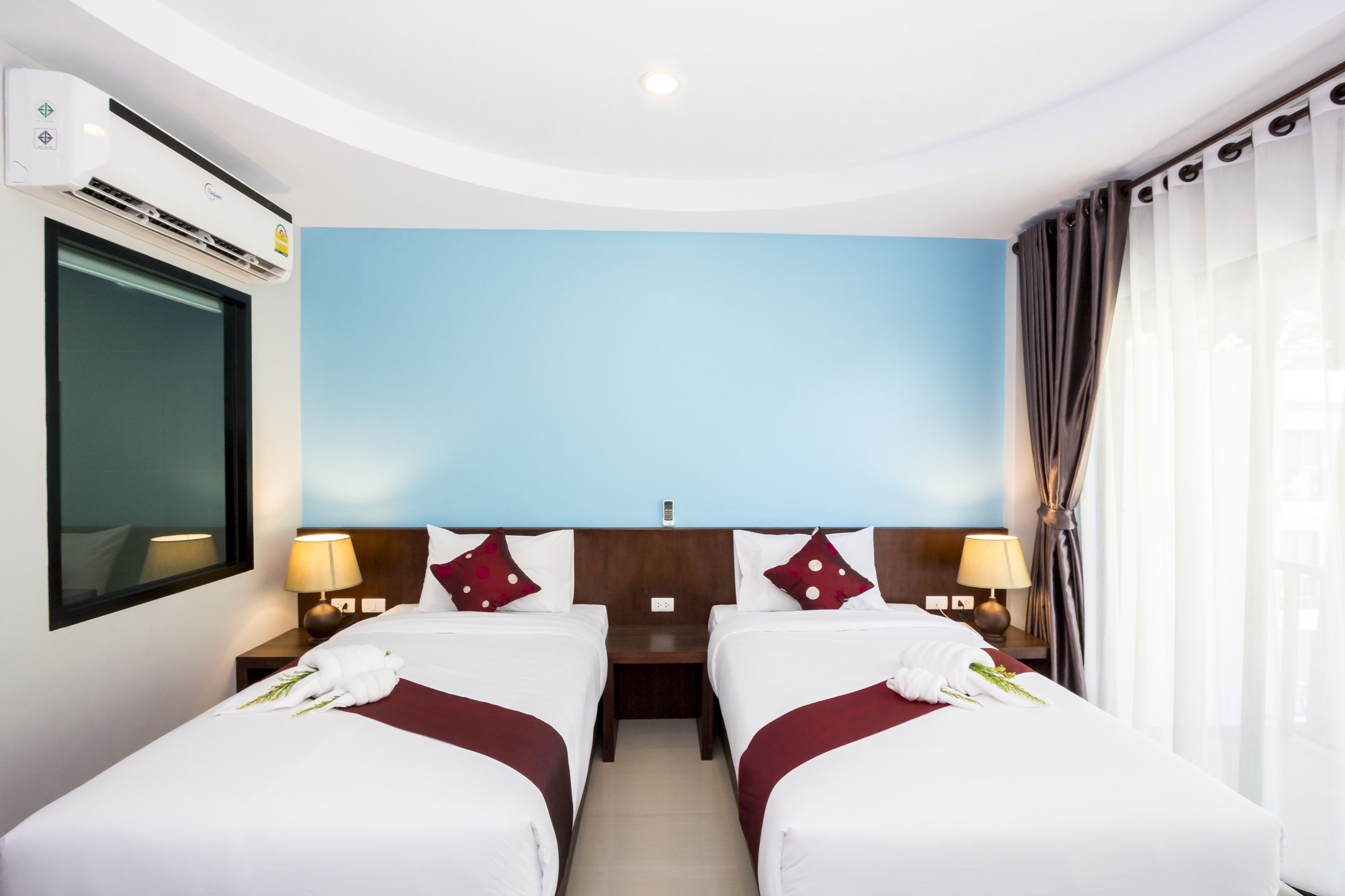 Una habitación de hotel con dos camas, un ventanal con cortinas, un aire acondicionado de pared y una mesita de noche con lámpara.