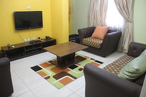 A living room with a flat-screen TV mounted on the wall, a wooden coffee table, two sofas with patterned cushions, and a colorful rug. - Magmercy Apartments (Lagos)
