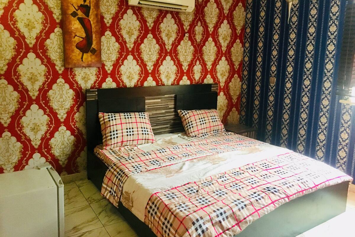 A bedroom with a bed, a headboard, a wall-mounted air conditioner, and patterned wallpaper.