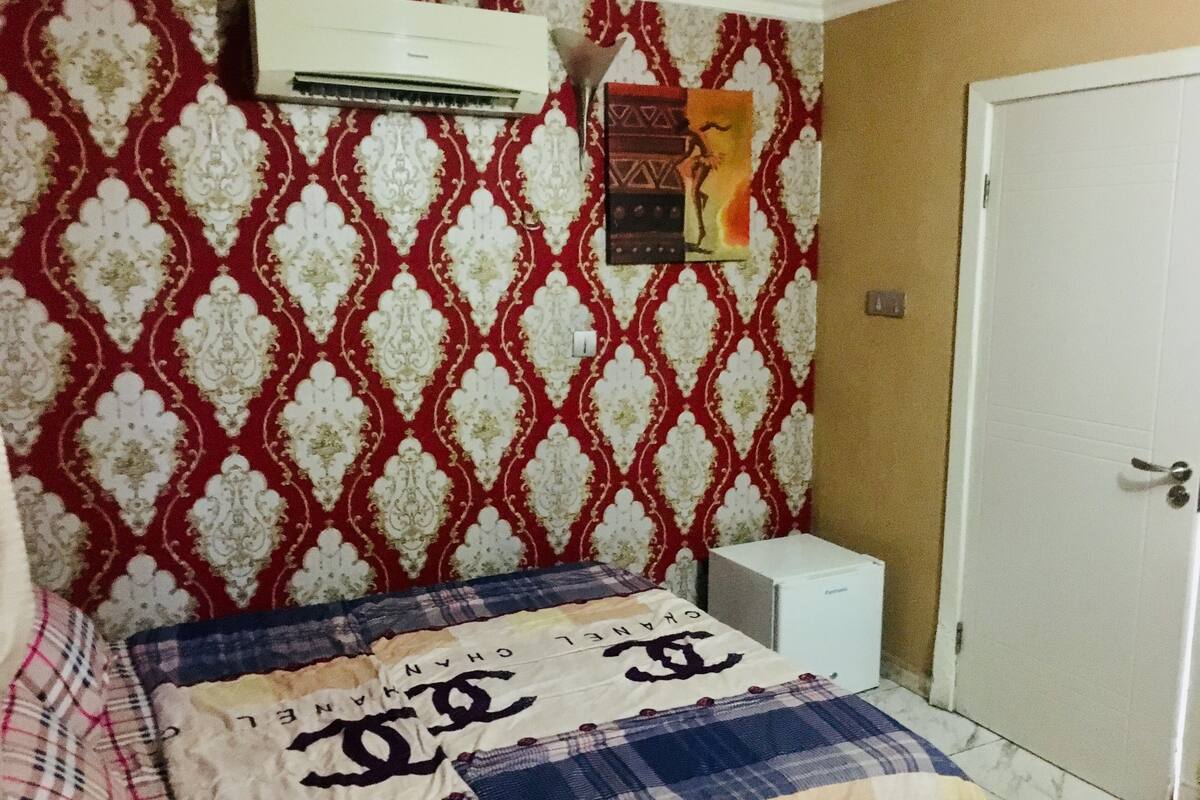 A bedroom with a patterned wallpaper, a bed with a blanket featuring a brand logo, a small white bedside table, and an air conditioning unit on the wall.