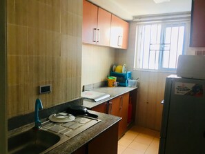 A kitchen with a sink, countertop, and cabinets. There is a window with bars and a door. - Magmercy Apartments (Lagos)