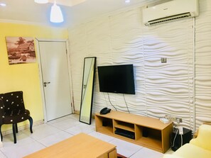 A living room with a flat-screen TV mounted on a textured wall, a wooden coffee table, a sofa, a chair, and a door. - Magmercy Apartments (Lagos)