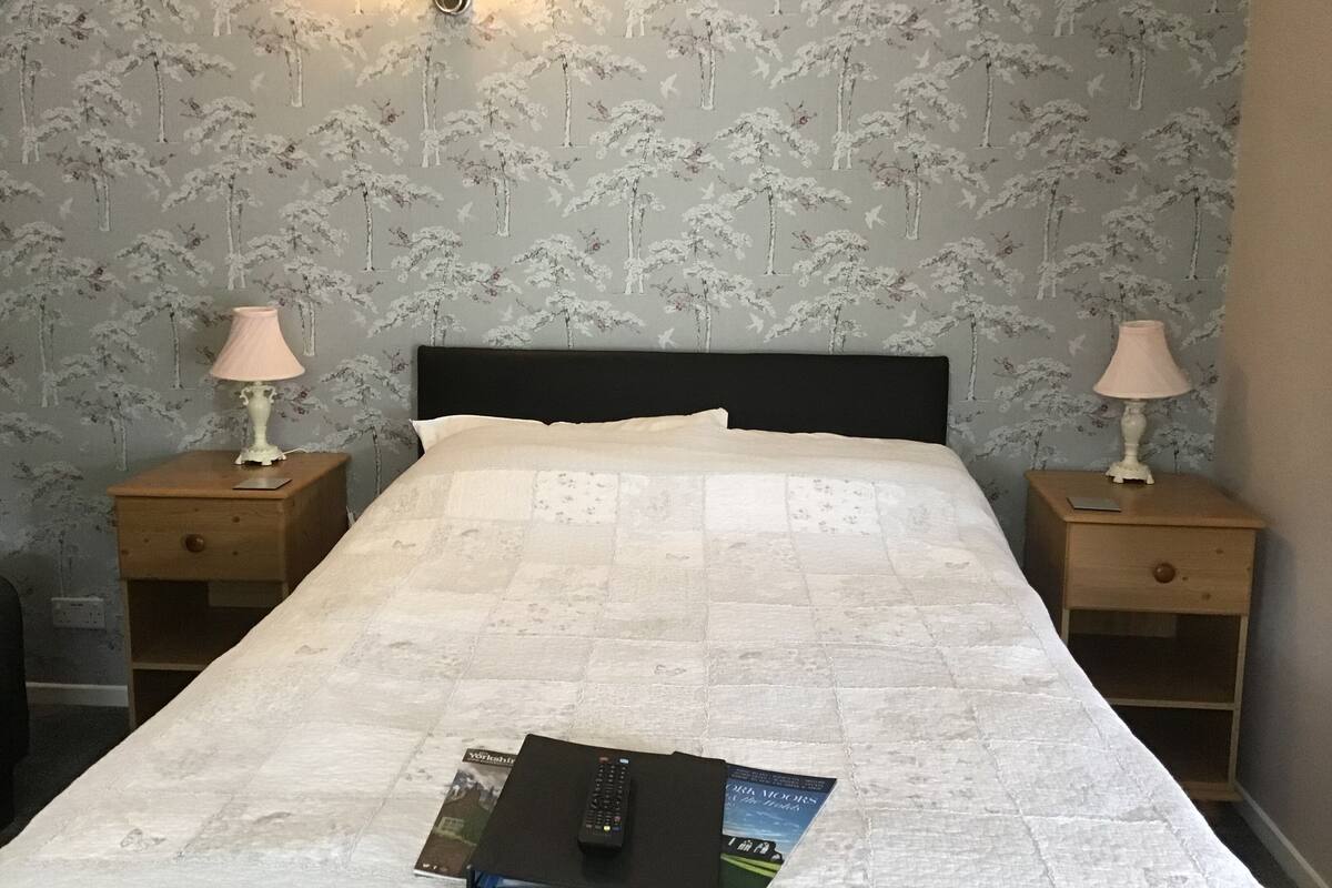 A neatly made bed with a quilt, two bedside tables with lamps, a wall-mounted light, and a remote control on the bed.