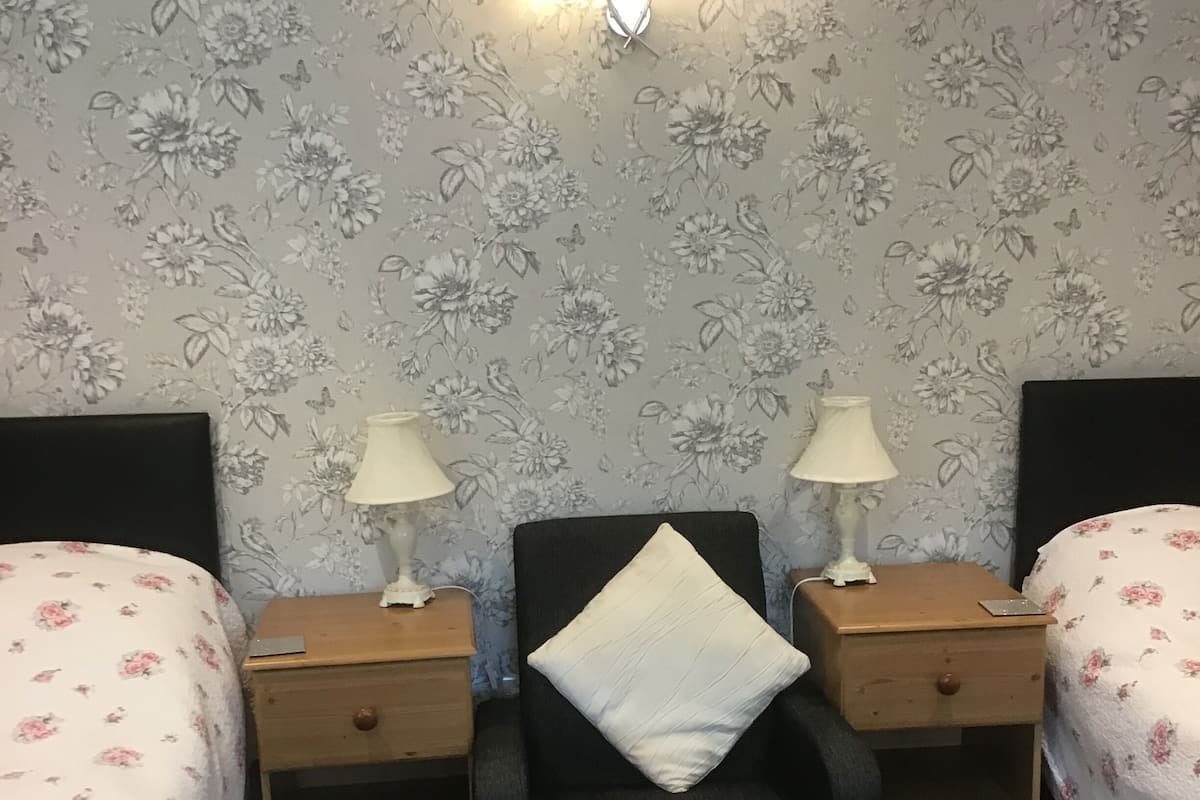 A bedroom with floral wallpaper, two bedside tables with lamps, a black chair with a pillow, and a bed with floral bedding.