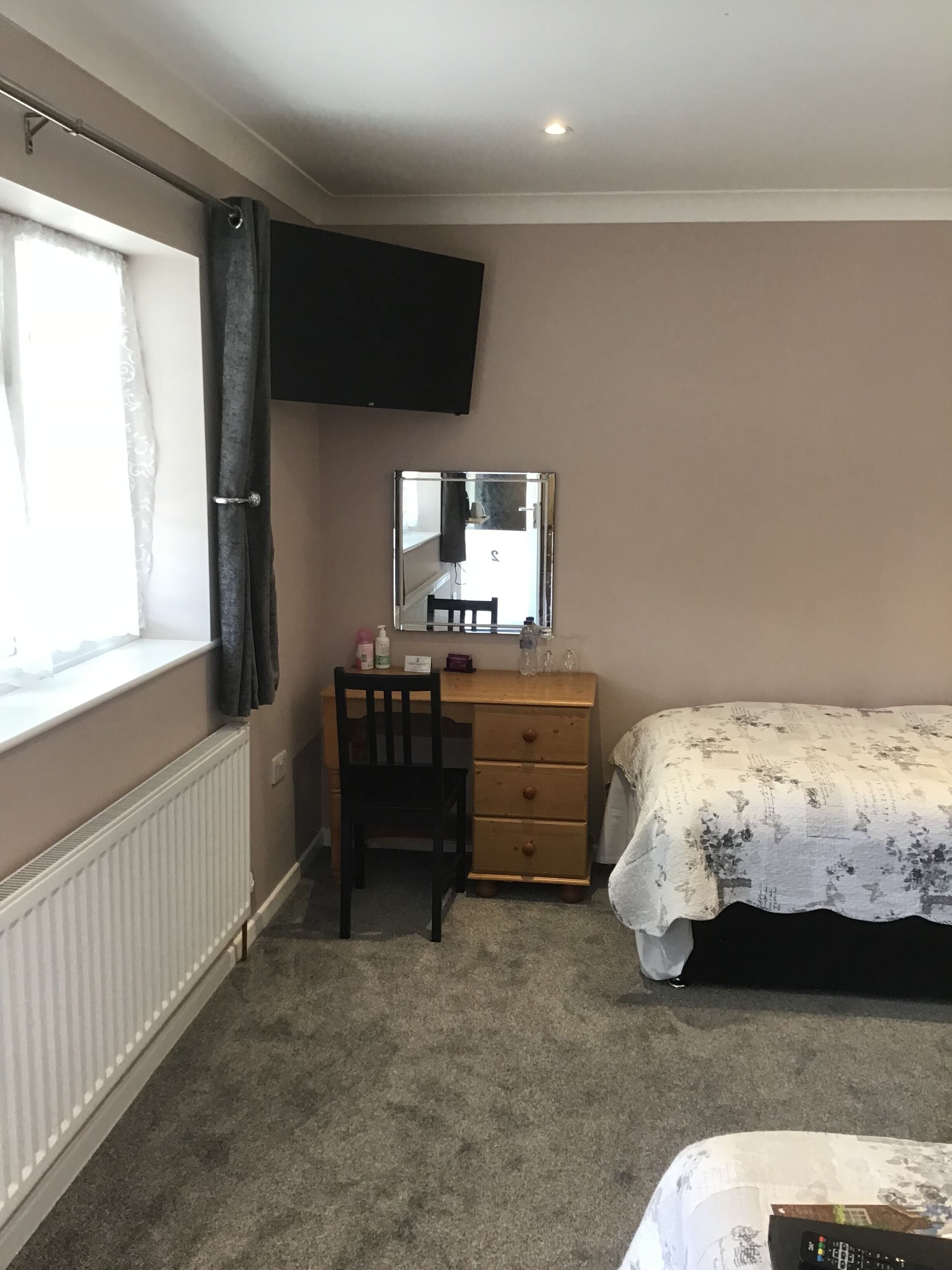Un dormitorio con cama, escritorio con silla, cómoda, espejo y ventana con cortinas.