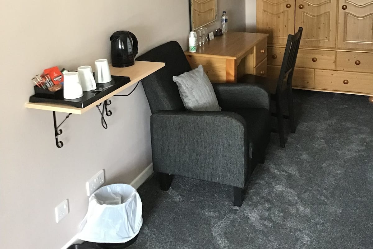 A compact hotel room with a single grey armchair, a small wooden desk, a mounted flat-screen TV, and a wardrobe with a mirror.