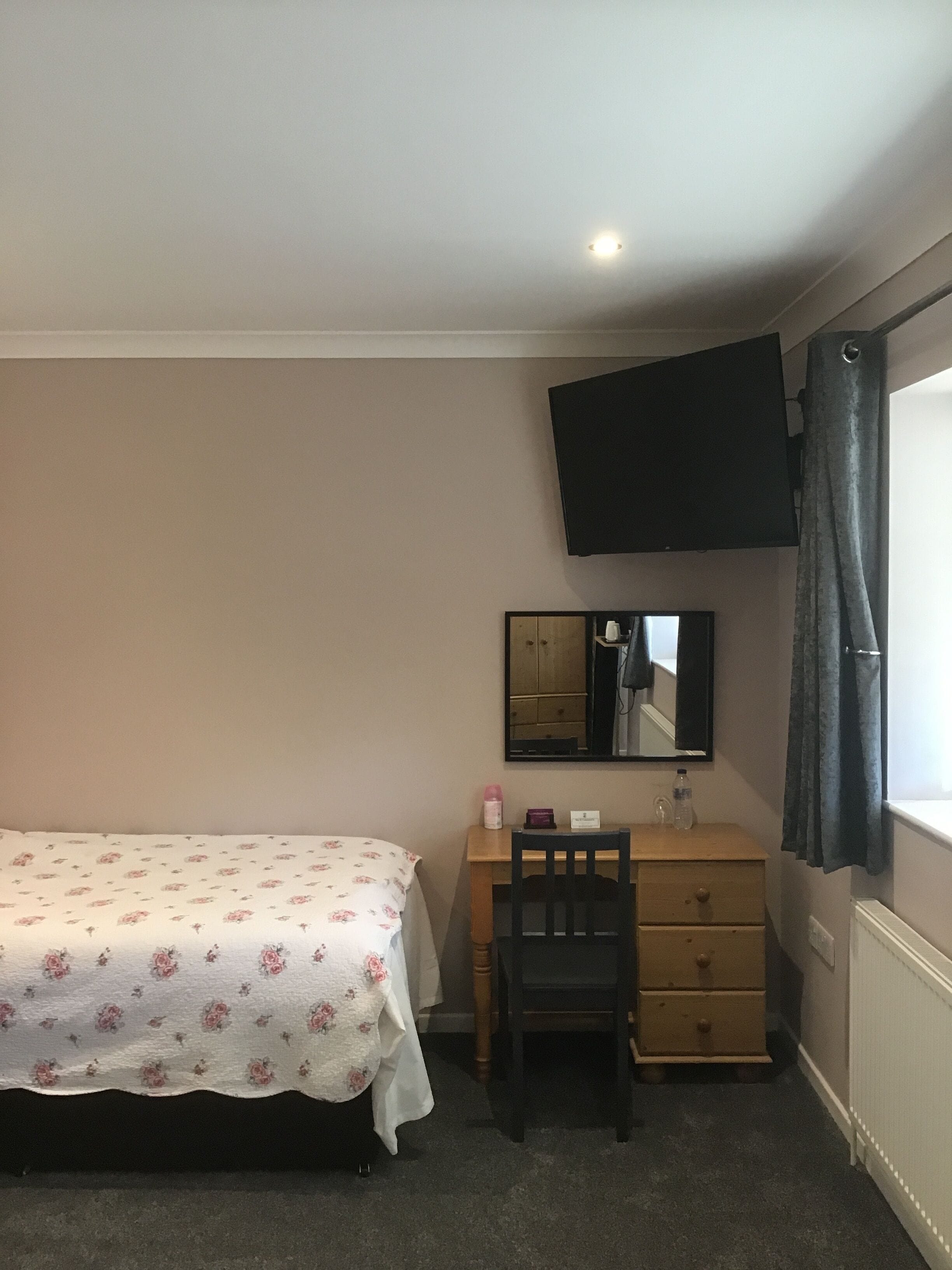 Un dormitorio con cama, escritorio, silla, cómoda, espejo y televisor montado en la pared.