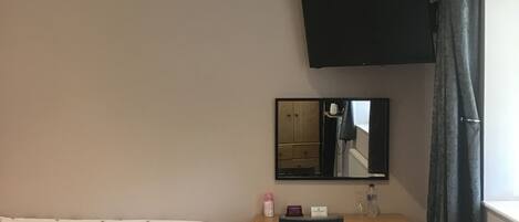 Un dormitorio con cama, escritorio, silla, cómoda, espejo y televisor montado en la pared.