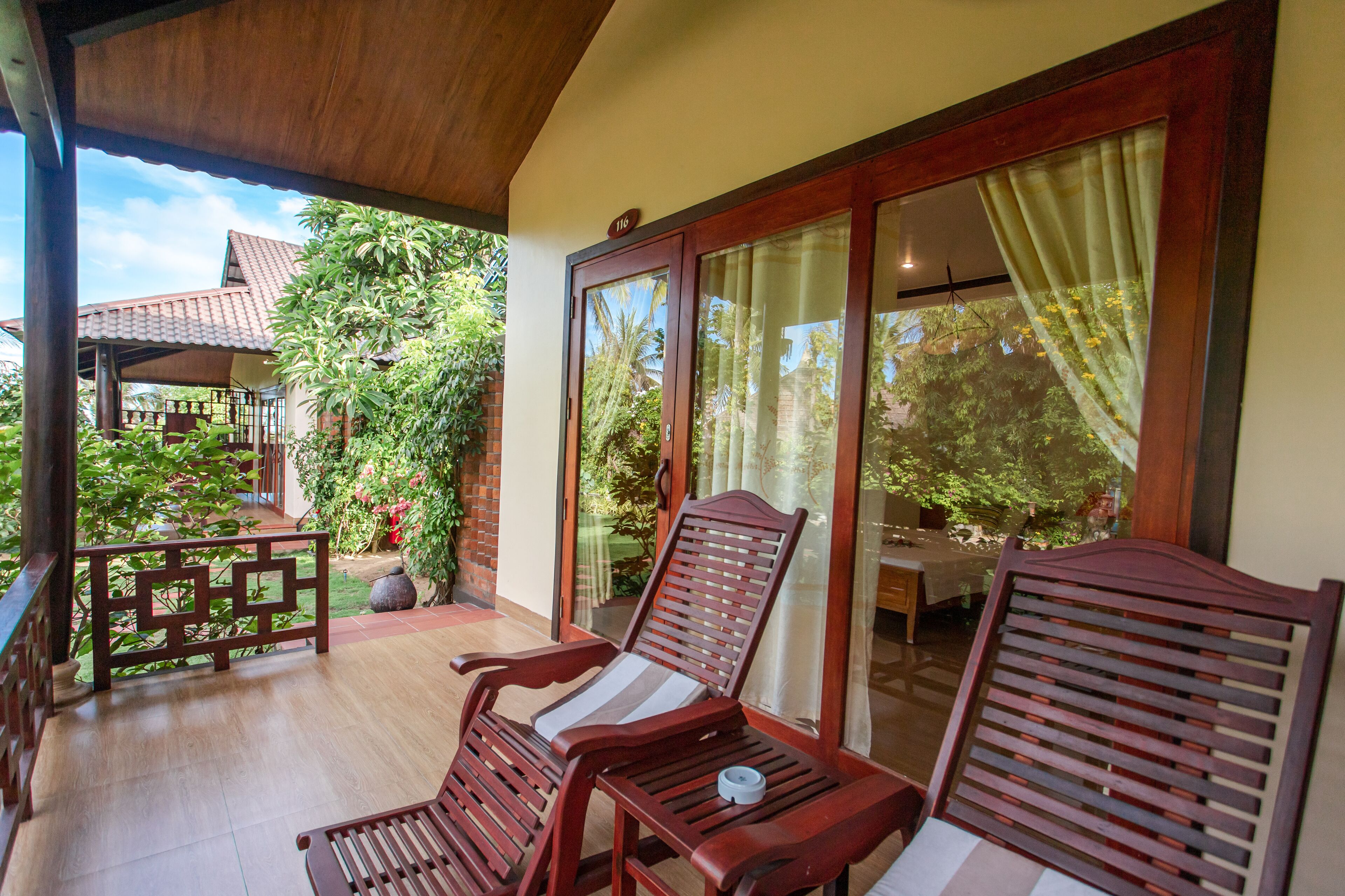 A covered balcony with wooden furniture, a glass door, and a view of a garden and building.