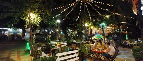 Un área de comedor al aire libre con mesas y sillas, rodeada de follaje e iluminación con luces colgantes.