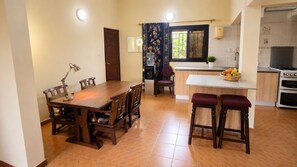 A dining area with a wooden table and chairs, a kitchen with a stove and oven, and a window with curtains. - Roc Heights Lodge (Bakau)