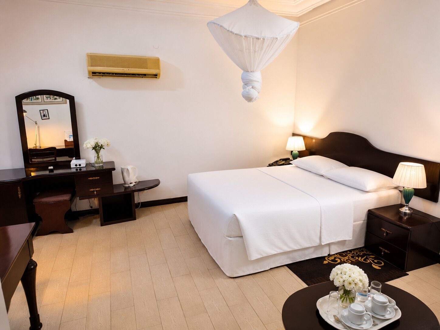 Quarto de hotel com cama, mesas de cabeceira, escrivaninha, cadeira, espelho, ar condicionado e um dossel decorativo.
