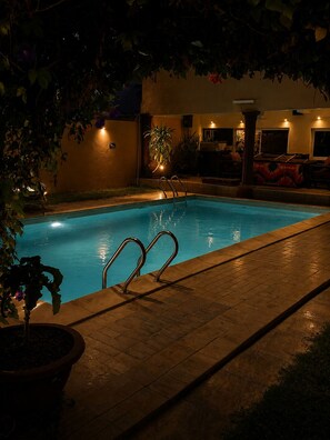 A swimming pool with metal ladders, surrounded by a tiled deck, potted plants, and a wall with a door and windows.
