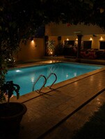 Ein Swimmingpool mit Metallleitern, umgeben von einer gefliesten Terrasse, Blumentöpfen und einer Mauer mit Tür und Fenstern.