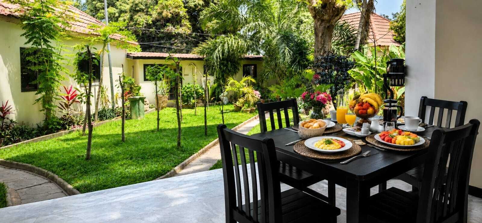An outdoor dining area with a table set for breakfast, surrounded by lush greenery and a small building in the background.