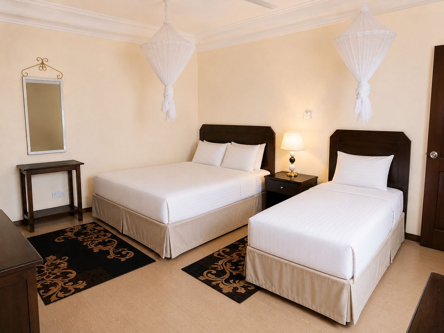 A hotel room with two single beds, a bedside table with a lamp, a mirror, and a decorative rug.