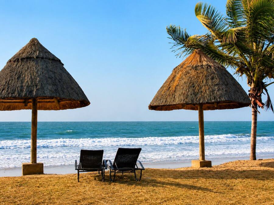 A beach with two thatched umbrellas, two lounge chairs, and a palm tree.
