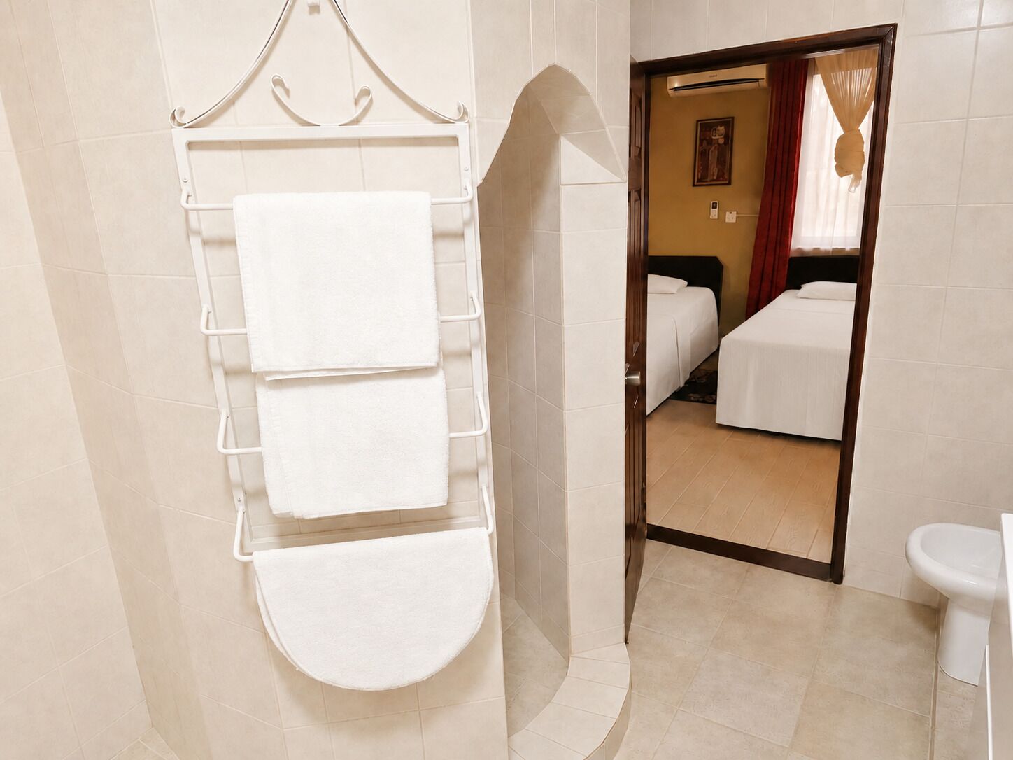 Un baño con un perchero que sostiene tres toallas, un inodoro y una cama visible a través de una puerta abierta.
