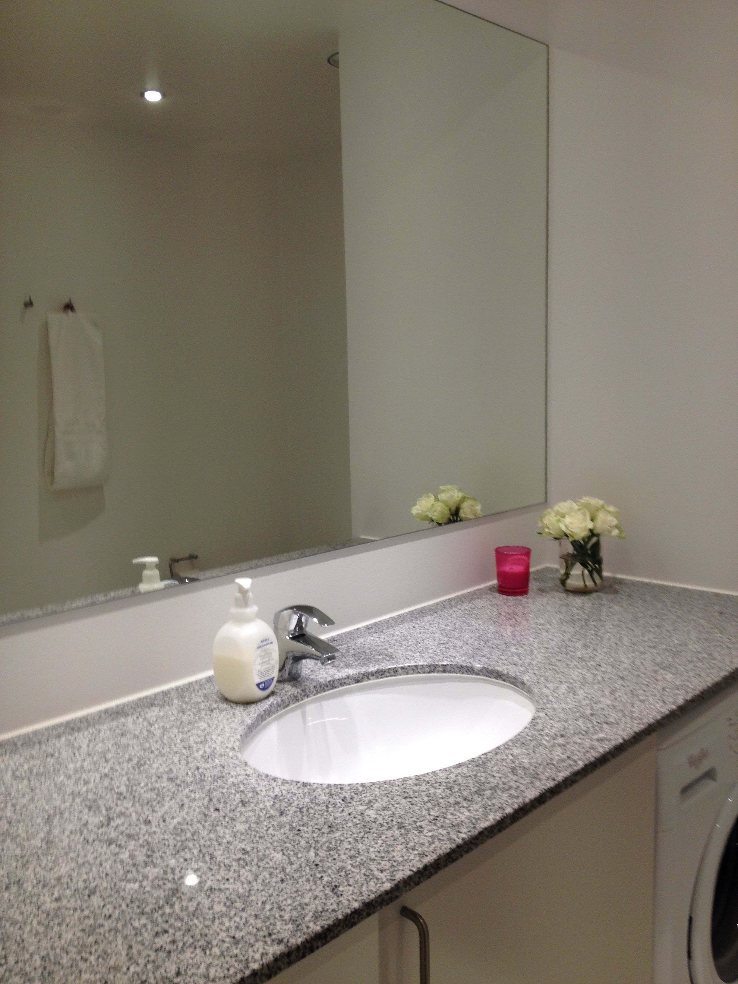 A bathroom with a granite countertop, a white sink, a mirror, a towel rack, a soap dispenser, a pink candle, and a vase with flowers.