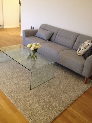 Un salon avec une table basse en verre, un canapé gris et un tapis beige.