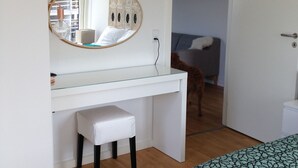 Un dormitorio con un escritorio blanco, un espejo redondo, un taburete pequeño, una cama con cubrecama estampado y una puerta que da a otra habitación.