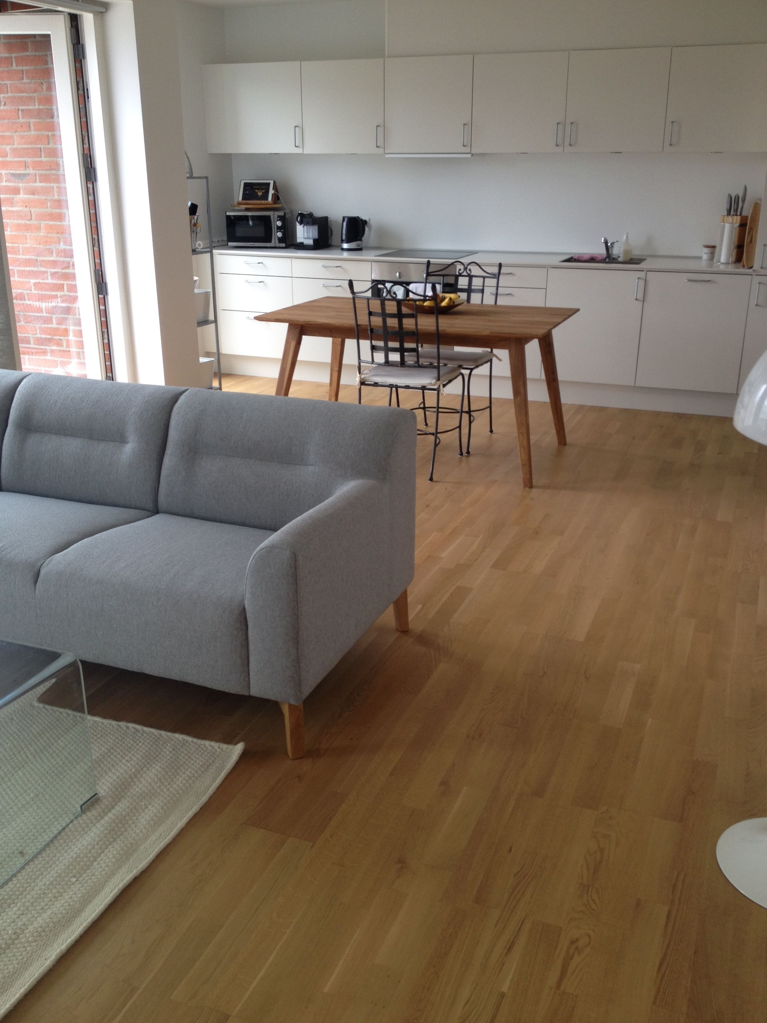 A modern living room with a grey sofa, wooden dining table, and chairs. A kitchen with white cabinets and appliances is visible in the background.