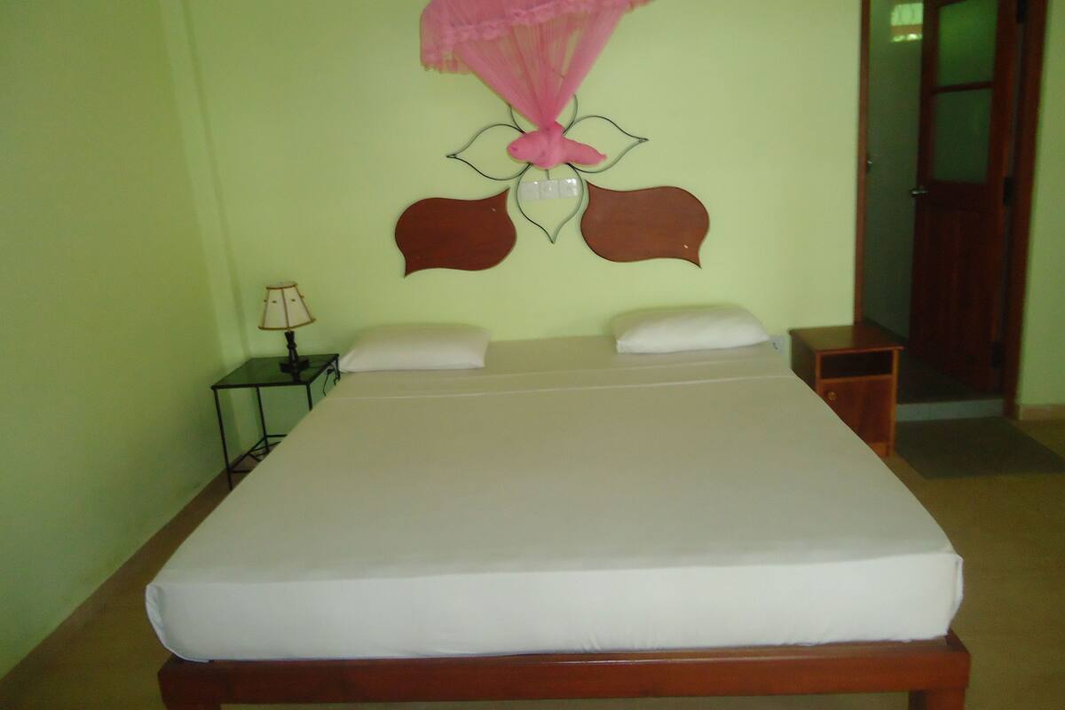 Una cama con colchón blanco, dos almohadas, una mesita de noche con lámpara y un adorno colgante sobre la cama.