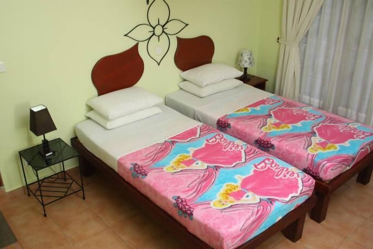 Una habitación con dos camas, una mesita de noche con lámpara, una decoración en la pared y una cortina.