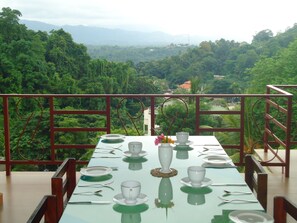 Una mesa de comedor dispuesta con platos, tazas y cubiertos, con vistas a un paisaje verde y exuberante con árboles y edificios a lo lejos.