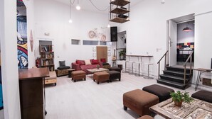 Un vaste espace de vie comprenant un ensemble de canapés, des poufs, une table basse et une bibliothèque. Un escalier mène à un niveau supérieur.
