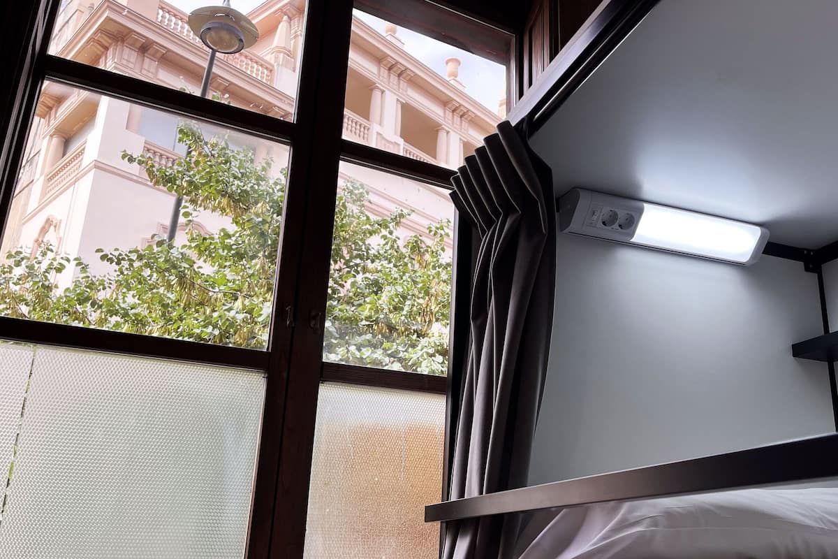 Ein Schlafzimmer mit Etagenbetten, einem Fenster, einer Deckenleuchte und Blick auf ein Gebäude mit Balkonen.