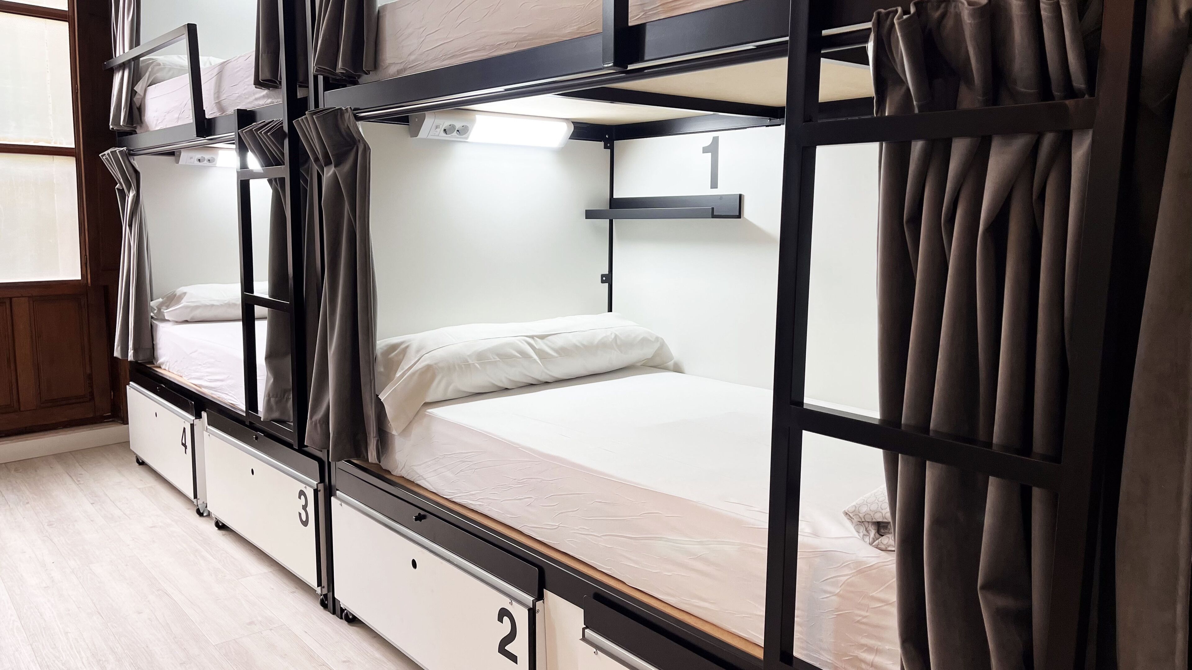 Camera con letto a castello, scrivania estraibile, finestra con tende e un numero sulla parete.