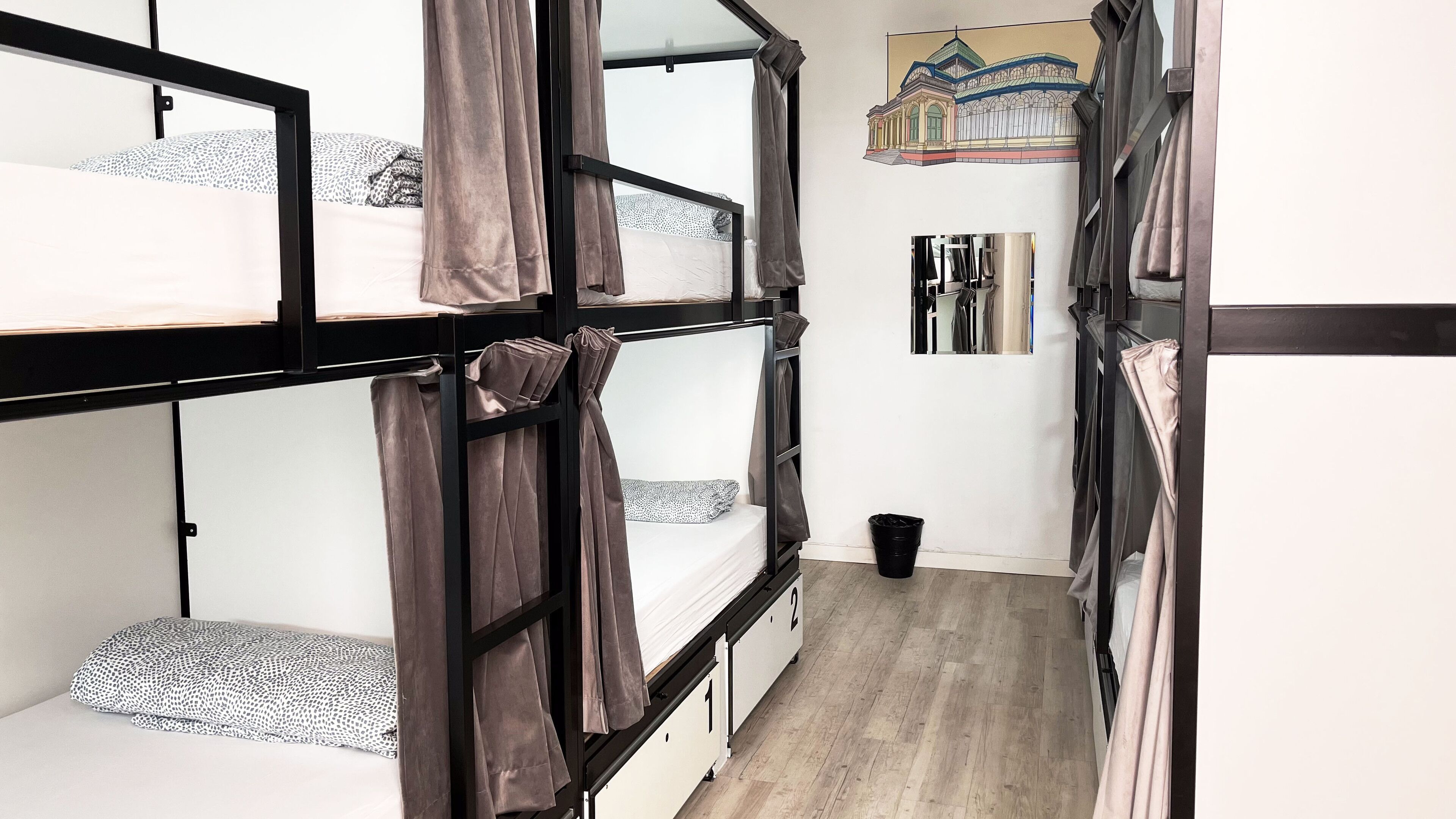 A dormitory room with bunk beds, a small trash bin, and a painting of a building on the wall.