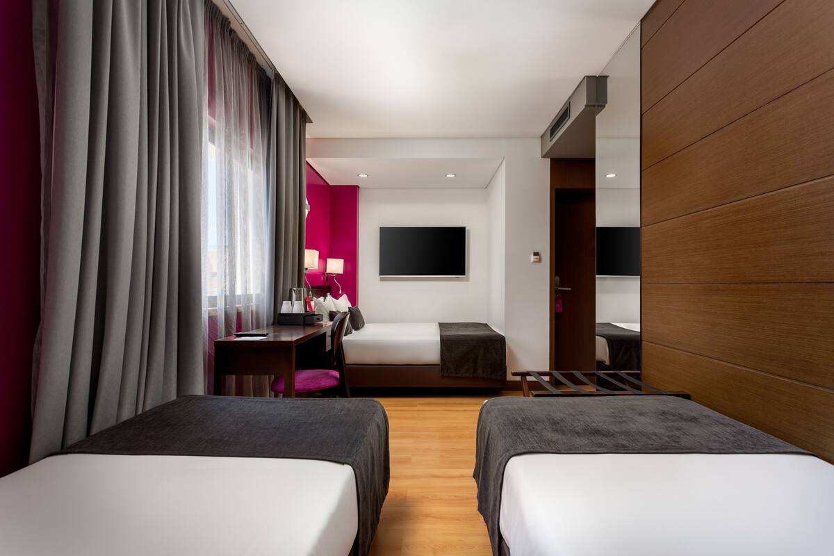 Ein Hotelzimmer mit zwei Betten, einem Schreibtisch, einem Stuhl, einem Fernseher und einem großen Fenster mit Vorhängen.