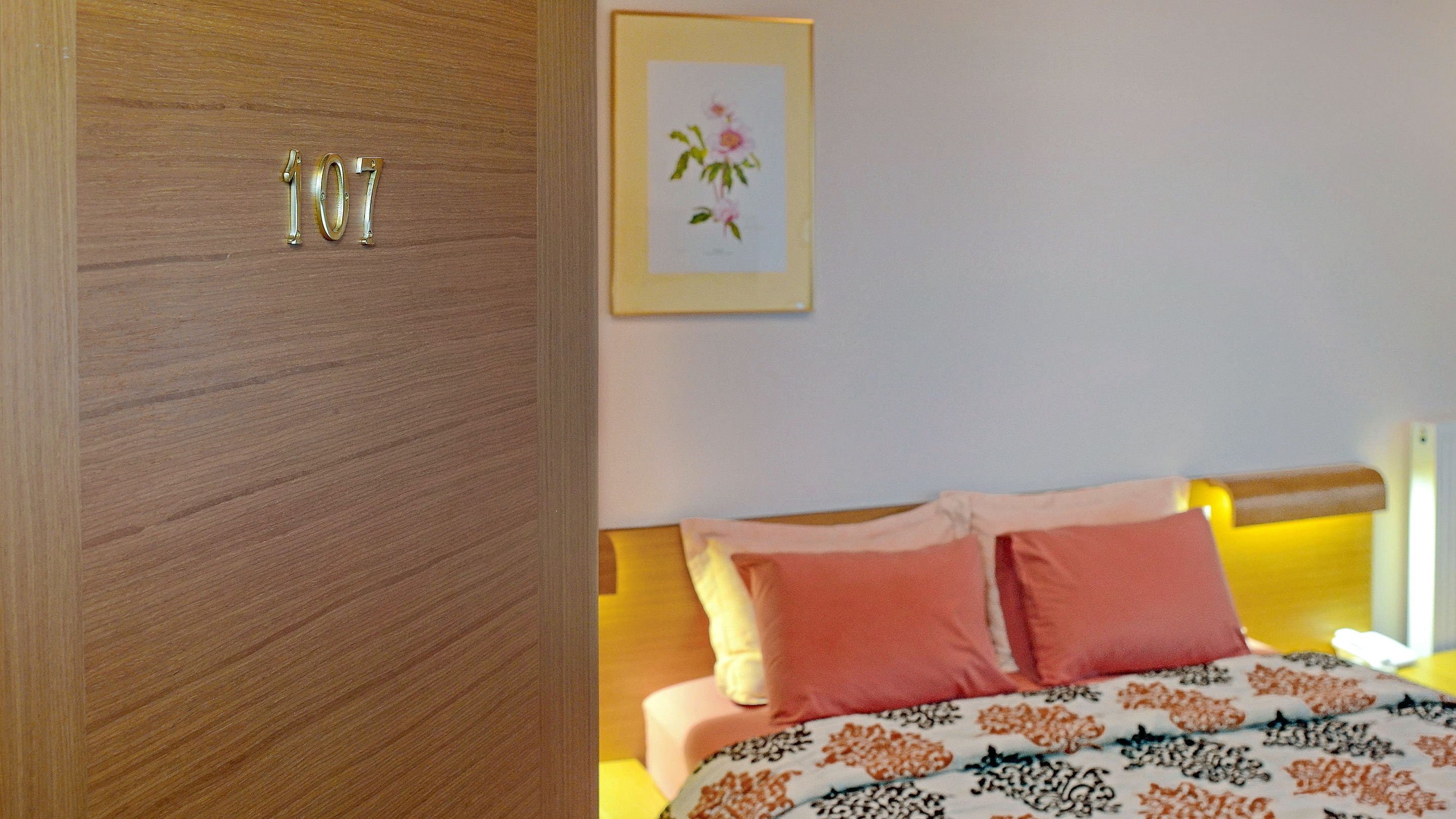Uma porta de madeira com o número 107, uma cama com roupa de cama rosa e branca e uma obra de arte floral emoldurada na parede.