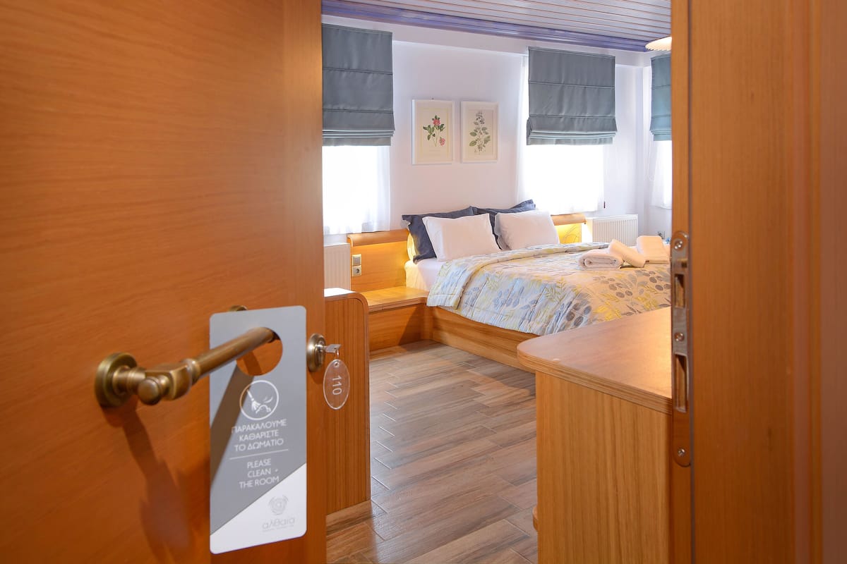 A hotel room with a wooden floor, a bed with a floral bedspread, two bedside tables with lamps, and a window with a view of the outside.