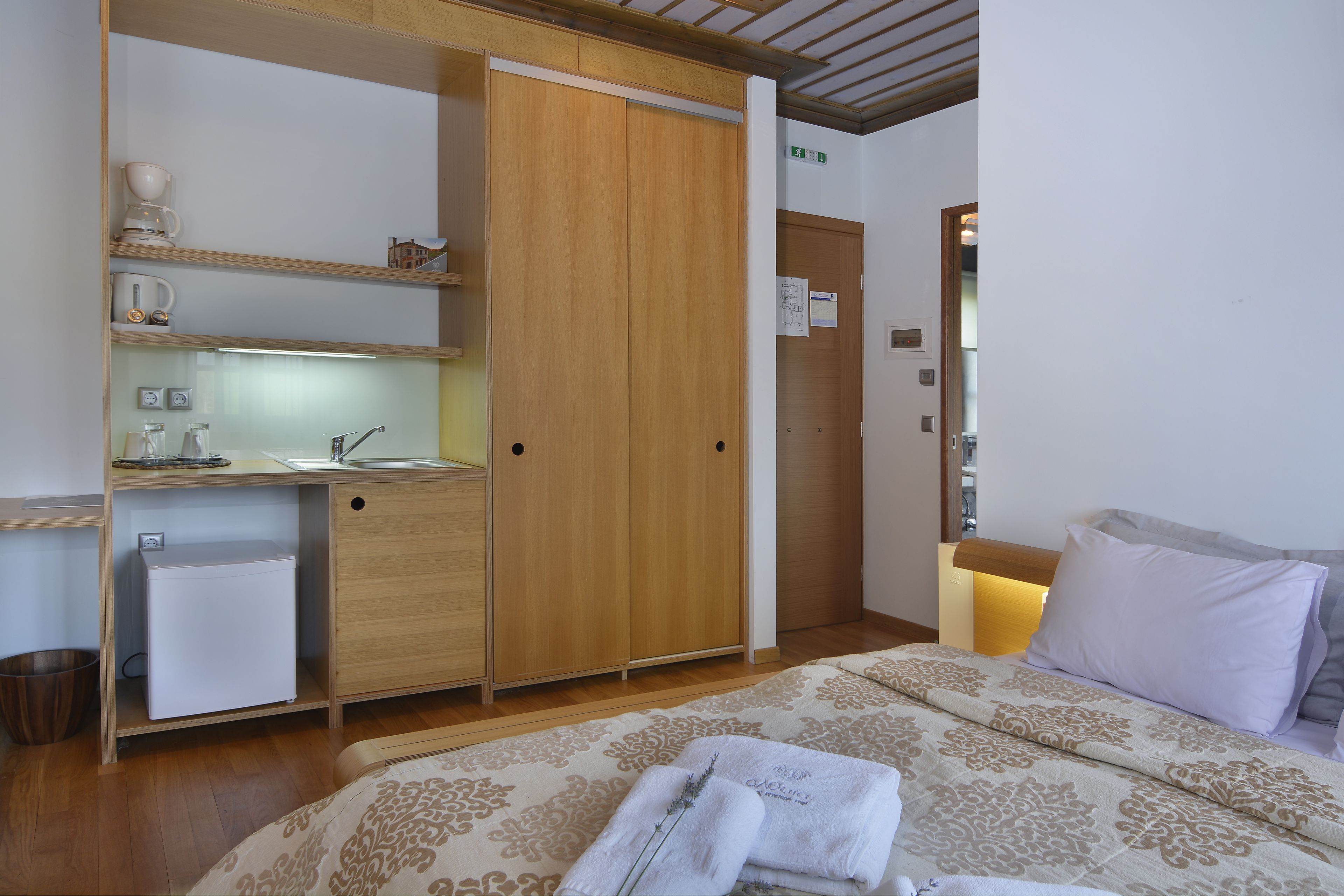 Una habitación de hotel con cama, un armario de madera, una pequeña kitchenette y una mesita de noche con lámpara.