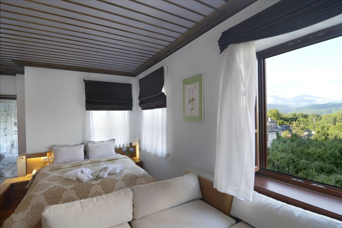 A bedroom with a large bed, a view of greenery and mountains, and a wooden ceiling.
