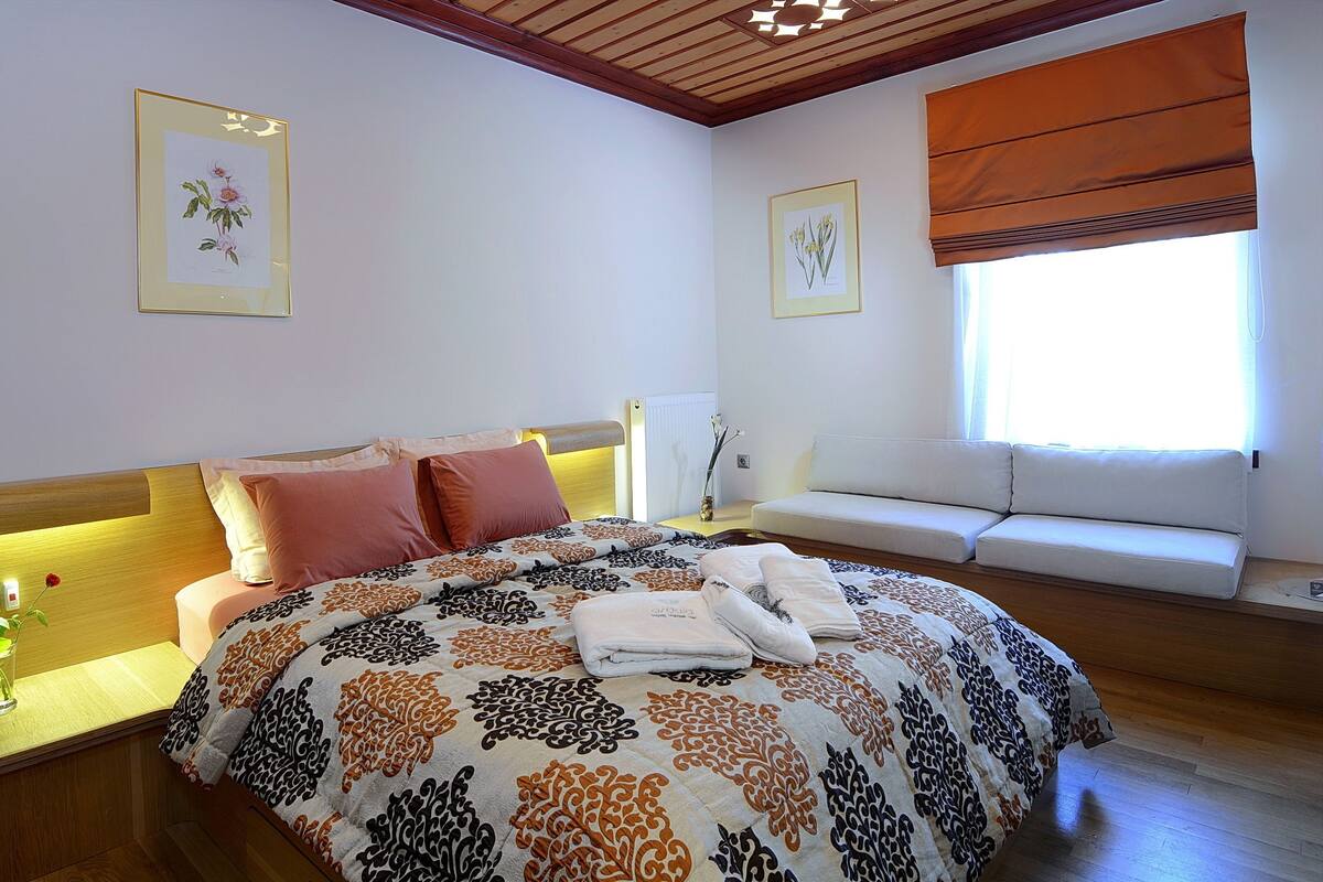 A bedroom with a bed, a bedside table with a vase of flowers, a window with blinds, and a wall-mounted radiator.