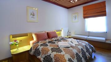 Un dormitorio con cama, una mesita de noche con un florero, una ventana con persianas y un radiador fijado a la pared.