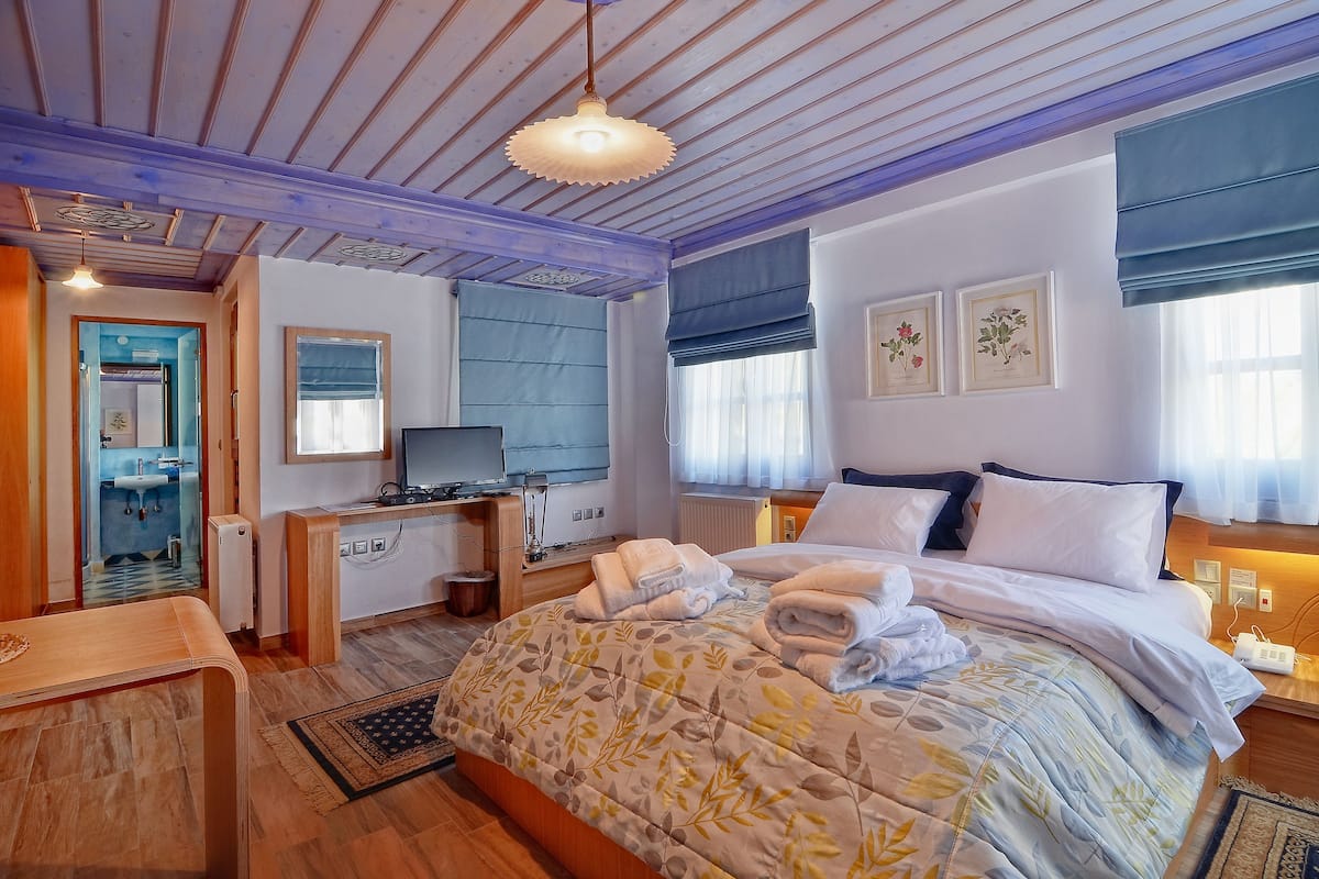 A hotel room with a bed, bedside table, desk with a computer, a mirror, a window with curtains, and a wall-mounted TV.